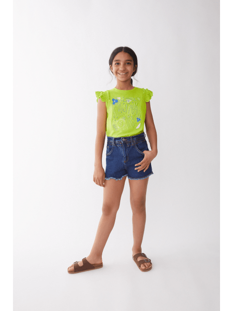 Niña sonriente modelando blusa verde limón Street Monkey con diseño "Limon" y shorts de mezclilla.